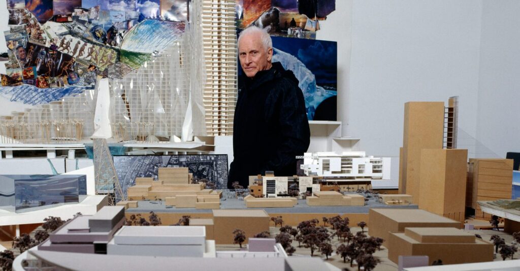 wide angle photograph of the architect, a white male with black shirt and pants surrounded by small scale building models